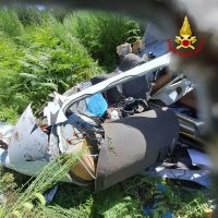 Auto parzialmente schiacciata tra erba alta, soccorritori sul posto, incidente stradale in area verde; tetto e paraurti distrutti.