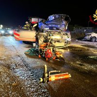 Auto incidentata di notte con veicoli fermati e soccorso sul ciglio della strada; cavi e strumenti da dettaglio caduti sull’asfalto bagnato.