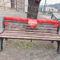 Panchina in legno con riflettori rossi e cuore rosso al centro, decorazioni laterali, in una piazza urbana.