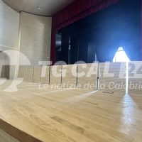 Scena di un palco vuoto con parquet chiaro, tende rosse e luce blu in fondo; testo sfumato sull’angolo presenta TTC...