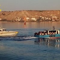 Barchetta sul mare calmo con costa rocciosa sullo sfondo e persone su una nave, luce dorata al tramonto in Italia.