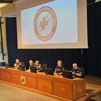Aula universitaria con panel di relatori seduti al tavolo, schermo proiettato sul logo circolare dell’istituzione, atmosfera formale.