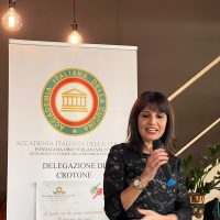 Donna sul palco che parla al microfono durante un evento formativo, con banner istituzionale sullo sfondo.