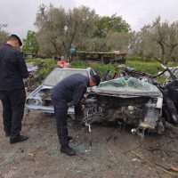 Soccorritori e polizia all’opera davanti a un’auto esplosa; danni frontali, incidenti in campagna, presenti personale sul posto.