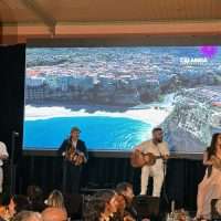 Band sul palco durante un evento, con pubblico presente e vista di mare sullo sfondo.