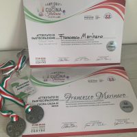 Premio certificato e medaglie in primo piano, confezione con loghi italiani e colori tricolore, celebrando una partecipazione culinaria.