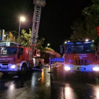 Auto antincendio e autobotti in strada bagnata di sera, soccorritori al lavoro e mezzi di emergenza accesi.