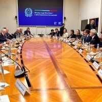 Sala riunioni ampia con lungo tavolo in legno, partecipanti in discussione, presentazione su schermo sullo sfondo.