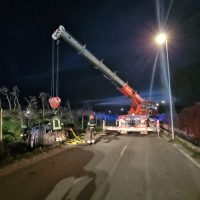 Lavoratori e gru in strada di notte, soccorso richiesto su veicolo incidentato, luci lampeggianti e presenza di strade chiuse.