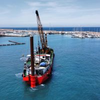 Veduta aerea di ormeggio marino: una gru su una barca rossa estrae materiali vicino al porto, acque azzurre e orizzonte marino.