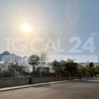 Strada urbana al tramonto, sole splendente su un muro decorato, alberi sullo sfondo e cielo azzurro.