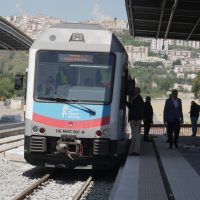 Treno in stazione metropolitana all'aperto, con passeggeri in attesa sul marciapiede, sfondo collinare e edificio storico.