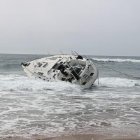 grande vela da trimarano incagliata sulla riva, onde morbide e mare grigio sullo sfondo, cielo coperto e sabbia bagnata.
