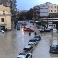 Strada allagata in una città italiana: veicoli parcheggiati e passanti attraversano grandi pozze d’acqua davanti a palazzi e negozi