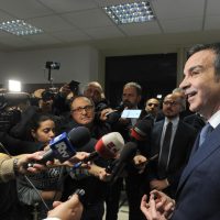 Uomo in completo blu parla tra cronisti e reporter in una sala conferenze, luci e microfoni puntati su di lui.