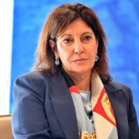 Donna professionista in primo piano, abbigliata in blu con sciarpa multicolore, sfondo blu, espressione seria e concentrata.