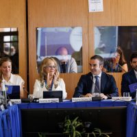 Un gruppo di professionisti seduti a un tavolo durante una conferenza, sullo sfondo pannelli e fotografie, moniti visibili.