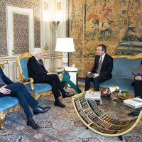 Quattro uomini seduti in salotto decorato, tavolino in vetro con libri, lampada e un grande tappeto, discussione formale.