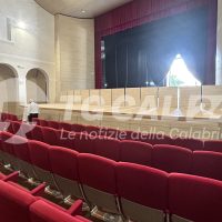 Sala teatrale con palcoscenico illuminato, poltrone rosse e pareti chiare, atmosfera elegante e organizzata per spettacolo.