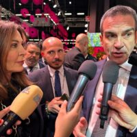 Intervistato da diverse TV mentre parla al microfono, circondato da intervistatori in un evento affollato e colorato.