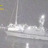 Imbarcazioni da pesca o motovedetta lucide in mare notturno, luci e riflessi sull’acqua, silhouette di vele e figure a bordo.