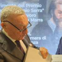 Anziano uomo in giacca e occhiali sfoglia un documento, sfondo blu con testo italiano di premiazione.