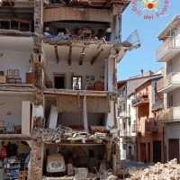 Edificio parzialmente crollato con detriti e strumenti da costruzione sparsi, segnaletiche e gente in zona; grida di soccorso in un contesto urbano.