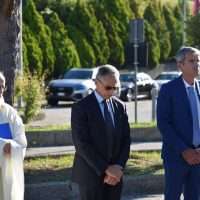 Tre uomini in passerella, due in completo blu e nero, una persona in abiti chiari, sfondo di alberi e veicoli in area esterna.