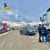 Auto parcheggiate su una strada fangosa tra rimorchi e camion, due persone parlano, cielo nuvoloso, atmosfera di cantiere o deposito stradale.