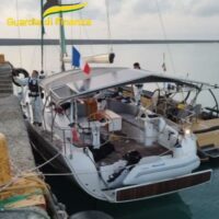 Barca a vela ormeggiata al molo con bandiere; riflessi d’acqua e cielo grigio sul porto, immobile tra giri di cordami e attrezzature.