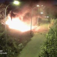 Explosione luminosa in strada al buio, fuoco alto e fiammecircondato da alberi e marciapiede, atmosfera di incidente notturno.