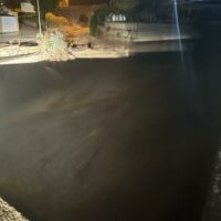 Lago calmo al tramonto circondato da rocce e un muro, riflessi neutri sull’acqua e una luce soffusa sulla sponda.