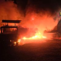 Macchina e fuoco intenso di notte, fiamme rosse avvolgono l’area, atmosfera tesa e fumo sullo sfondo.
