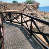Sentiero su passerella di legno lungo la scogliera: vista sul mare azzurro e su una costa rocciosa.