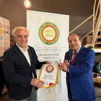 Due uomini in giacca stringono una targa tra logo e banner; gesto formale in ambiente professionale.