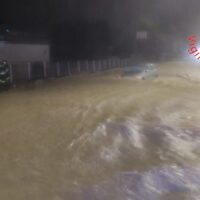 Strada allagata di notte con acque mosse, lampioni fiocchi di luce e edifici sullo sfondo.