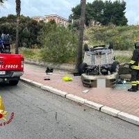 Incidente stradale: auto capovolta su marciapiede, soccorritori e ambulanza presenti, residente vicino osserva da lato.