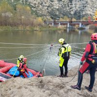 Personaggi in tuta di salvataggio su una riva di fiume, con corde e zaini, tra alberi in fiore e un pontile sullo sfondo.