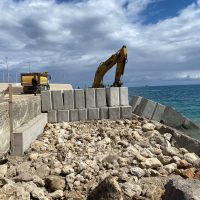 Molo in costruzione con escavatore giallo e blocchi di cemento, roccioso ancor più vicino al mare azzurro e cielo nuvoleggiante.