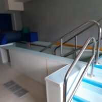 Piscina al chiuso con gradini azzurri, corrimano in metallo e area relax: spazio sportivo o termale, pareti neutre.
