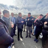 Gruppo di persone in abiti formali e militari si scambiano strette di mano all'aperto, sotto un cielo sereno.