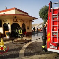 Immagine di un locale all’esterno con segnalazioni di emergenza, operativi in divisa camminano vicino a una scala rossa del furgone dei vigili del fuoco.