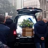 Auto con cofano aperto in corteo: bara bianca con fiori e urne, persone intorno in abiti scuri, in un contesto urbano.
