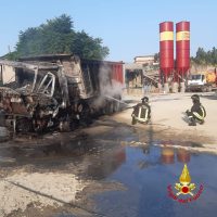 Incidente stradale con veicolo pesante semicarbonizzato, due soccorritori in abiti riflettenti accanto a pozza d’acqua e fiamme, logo in basso a destra.