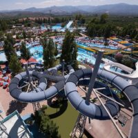 Complesso di piscine all'aperto con scivoli tubolari, lettini e ombrelloni, alberi e montagne sullo sfondo, atmosfera vacanziera.