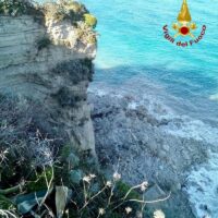 Scogliera rocciosa a picco sul mare azzurro con vegetazione mediterranea in primo piano e acque turchesi sullo sfondo.