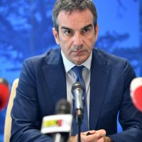 Uomo in smoking blu a conferenza stampa, fronte seria, microfono davanti, sfondo azzurro e sedie rosse sullo sfondo.