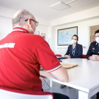 Gruppo di uomini in una riunione in ufficio: due presenti seduti al tavolo, uno in camicia, tutti con mascherine e un paio di visi non visibili.