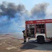 Vigili del fuoco in azione vicino a un'auto sullo sfondo, con fumo visibile e estintore collegato a una pompa, atmosfera tesa.