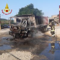 Camion incendiato irrorato da fiamme in piazzale, vigili del fuoco vicino a soccorsi, fumo e detriti sparsi, luce diurna, strade bagnate.